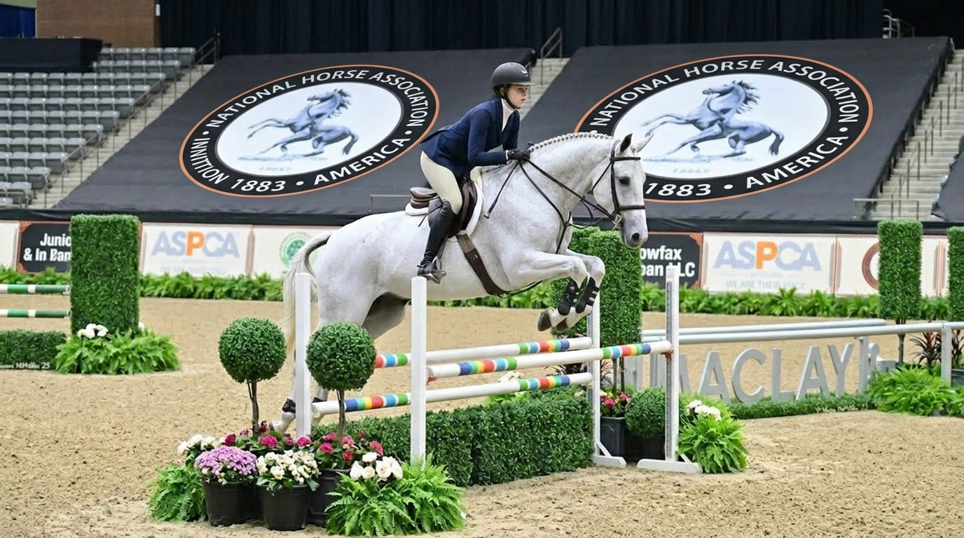 Junior rider on a grey warmblood horse mid-jump at the ASPCA Maclay National Championship, indoor arena, light tan breeches and black Samshield helmet, MACLAY signage visible on the arena floor — Notting Hill Equine
