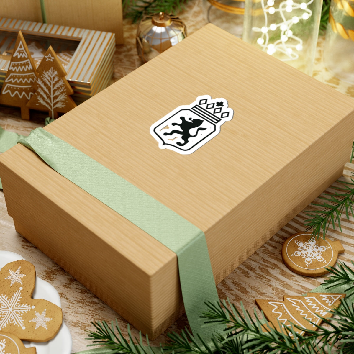 Gift box with a green ribbon and decorative logo on a festive background with cookies and lights.