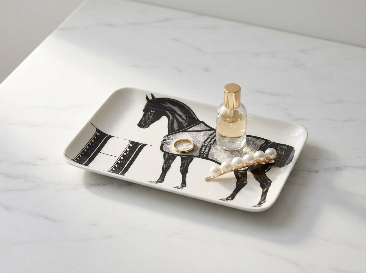 Ceramic equestrian catch-all tray with black horse motif styled on marble surface with gold ring, glass perfume bottle, and pearl hair clip